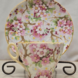 May include: A vintage teacup and saucer set. The cup and saucer feature a floral design with pink blossoms, brown branches, and green leaves on a cream backdrop. The edges are trimmed with gold. The set is displayed on a black metal stand.