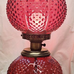 Może przedstawiać: Rubinowa szklana lampa typu hurricane z fakturą w kropki na kulistej podstawie i kloszu. Lampa posiada falbankę na górnej krawędzi klosza, mosiężną metalową podstawę oraz centralny mosiężny kołnierz z przełącznikiem obrotowym.