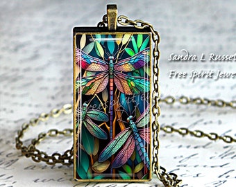 Puede incluir: Un collar con colgante rectangular con un diseño de libélula estilo vidriera. El colgante presenta una gran libélula central con alas coloridas y dos libélulas más pequeñas. La cadena es de color bronce y el texto "sandra L Russell Free spirit jewelry" es visible.