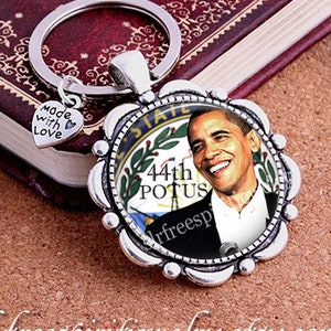 Puede incluir: Llavero plateado con un colgante circular con un retrato de Barack Obama, el 44º POTUS. El colgante tiene un borde festoneado y un pequeño dije de corazón con la inscripción "made with love".