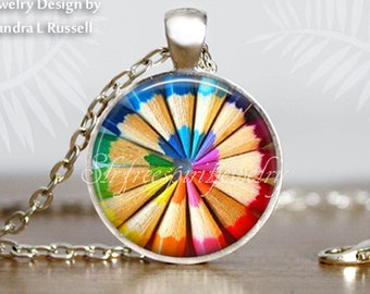 May include: A silver-toned necklace featuring a round pendant with a colorful pencil design. The pendant showcases a radial arrangement of pencil tips in various colors, including blue, green, yellow, orange, and red. The text "Jewelry Design by Sandra L Russell" is visible.