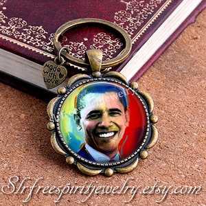Puede incluir: Un llavero de color bronce con un retrato de Barack Obama. La imagen está enmarcada en un marco decorativo festoneado. Un pequeño dije en forma de corazón con las palabras "Made with Love" cuelga del llavero. El nombre de la tienda Etsy es visible.