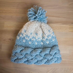 May include: A hand-knitted winter hat in light blue and white. The hat features a braided cable knit brim, a patterned design in the middle, and a fluffy pom-pom on top. The hat is made of soft yarn and is perfect for cold weather.