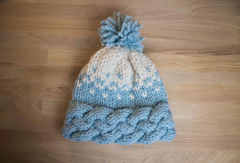 May include: A hand-knitted winter hat in light blue and white. The hat features a braided cable knit brim, a patterned design in the middle, and a fluffy pom-pom on top. The hat is made of soft yarn and is perfect for cold weather.