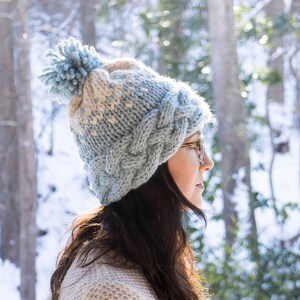 May include: A light blue and cream-colored cable-knit hat with a matching pom-pom. The hat is worn by a person with dark hair and glasses, set against a snowy, wooded backdrop. A cozy winter accessory.
