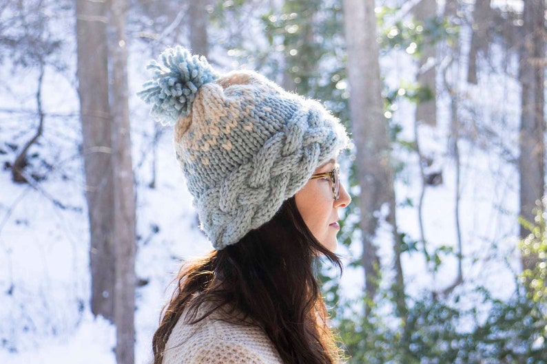 May include: A light blue and cream-colored cable-knit hat with a matching pom-pom. The hat is worn by a person with dark hair and glasses, set against a snowy, wooded backdrop. A cozy winter accessory.