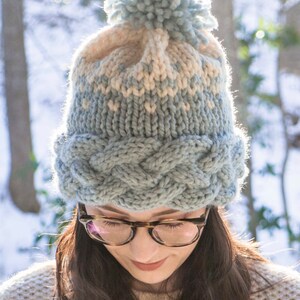 May include: A knitted winter hat with a light blue and cream color scheme. The hat features a braided cable knit band and a fluffy pom-pom on top. The hat is worn by a person with dark hair and glasses.