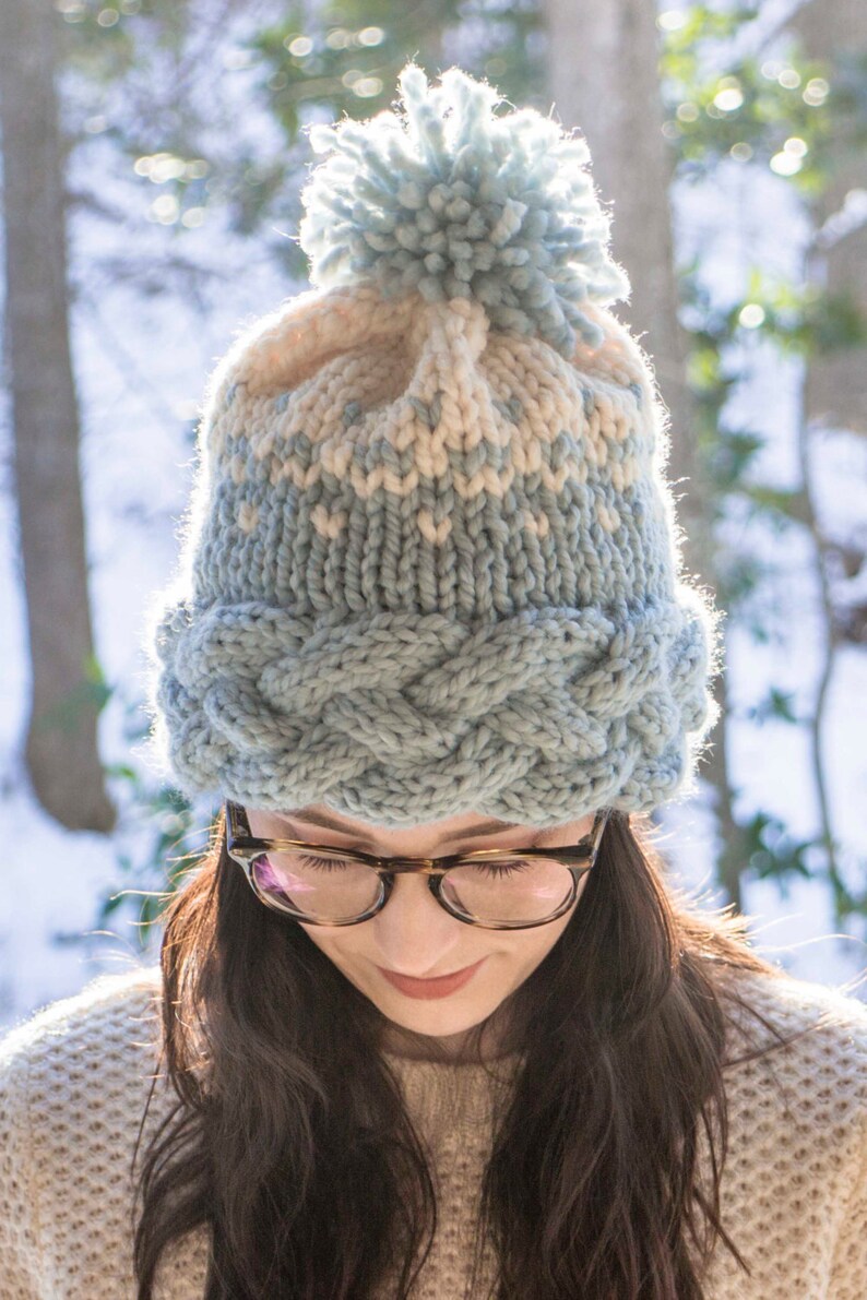 May include: A knitted winter hat with a light blue and cream color scheme. The hat features a braided cable knit band and a fluffy pom-pom on top. The hat is worn by a person with dark hair and glasses.