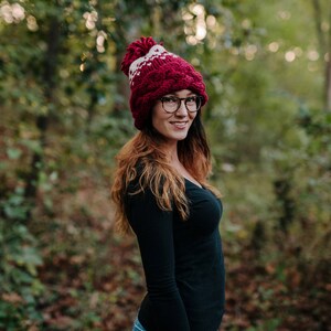May include: A burgundy and white knit hat with a pom-pom. The hat has a cable knit design and a white geometric pattern. The person is wearing black-framed glasses and a black long-sleeve shirt. The background is a blurred forest.