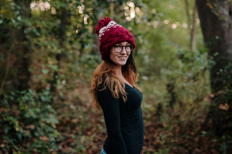 May include: A burgundy and white knit hat with a pom-pom. The hat has a cable knit design and a white geometric pattern. The person is wearing black-framed glasses and a black long-sleeve shirt. The background is a blurred forest.