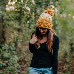 May include: A mustard yellow and white knit hat with a braided brim and a pom-pom on top. The hat is worn by a person wearing a black long-sleeve shirt and blue jeans. The background is a blurred forest scene.