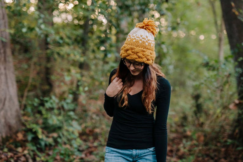 May include: A mustard yellow and white knit hat with a braided brim and a pom-pom on top. The hat is worn by a person wearing a black long-sleeve shirt and blue jeans. The background is a blurred forest scene.