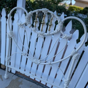 May include: Off-white metal bed frame with ornate detailing. The headboard features decorative scrollwork and vertical brass-colored accents. The bed frame is set against a white picket fence, with a blurred background of greenery and a clear sky.