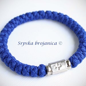 Puede incluir: Un brazalete de oración azul anudado con un dije de cruz de plata. El brazalete está hecho de cordón trenzado y tiene un cierre de plata. El texto "Srpska brojanica ©" es visible en el fondo.
