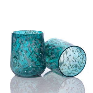 May include: Two stemless drinking glasses made of translucent teal glass. Each glass features a distinct mottled pattern of white and dark blue flecks embedded within the glass material, creating a textured appearance across the rounded surfaces.