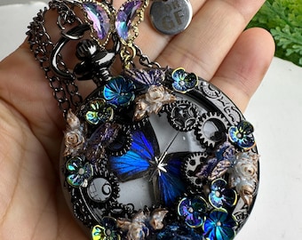 May include: A black, round pendant necklace with a butterfly design. The pendant features a blue butterfly, gears, and iridescent flowers. The necklace has a black chain and a small, round tag with the letters "GF".