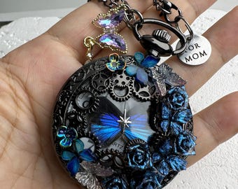 May include: A black steampunk-style pendant necklace with a round clock face design. The pendant features blue butterfly and rose accents, with a chain and a charm that says "FOR MOM".