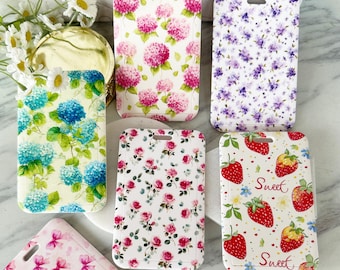 Puede incluir: Una colección de fundas rectangulares para teléfonos con diseños florales y de frutas. Las fundas presentan patrones de hortensias rosas, flores moradas, rosas rosas y fresas con la palabra "Sweet". Cada funda tiene un pequeño agujero en la parte superior.