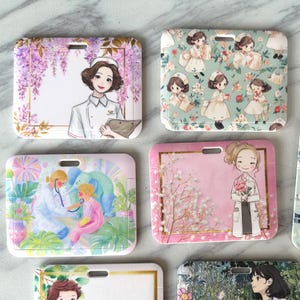 May include: A collection of rectangular badge holders with various illustrations. Designs include a nurse with wisteria, a doctor examining a child, and a floral pattern with cartoon characters. Each holder has a slot for a lanyard.