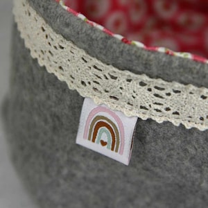 May include: A gray felt storage basket with a white lace trim and a pink and brown rainbow label with a heart in the center.