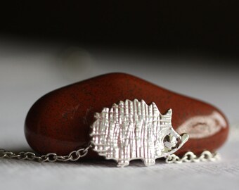 Könnte beinhalten: Halskette mit einem silbernen Anhänger in Form eines Igels. Der Anhänger ist ein detailliertes, strukturiertes Design eines Igels. Die Halskette ist eine zarte Silberkette. Der Anhänger liegt auf einem glatten, rotbraunen Stein.