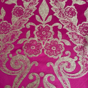 May include: Close-up of a vibrant fuchsia fabric with a repeating floral pattern in a metallic gold color. The design features stylized flowers, leaves, and swirling scrollwork, creating a luxurious and ornate texture.
