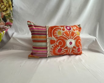 May include: A rectangular pillow with a white background and orange, pink, and yellow embroidered floral designs. The pillow has a striped fabric backing with a white braided trim.