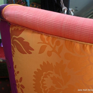 May include: Close-up of a section of a colorful upholstered chair. The chair features a vibrant orange floral patterned cushion, a salmon-colored top, and a purple side. The design includes a mix of textures and colors.