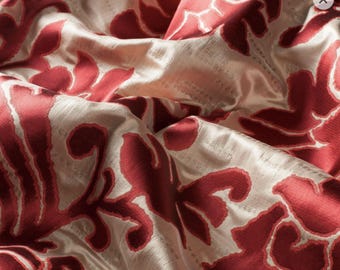 May include: Close-up of a luxurious fabric with a cream-colored background and a bold, repeating floral pattern in a deep red hue. The fabric has a soft sheen and a textured surface, suggesting a high-quality material.