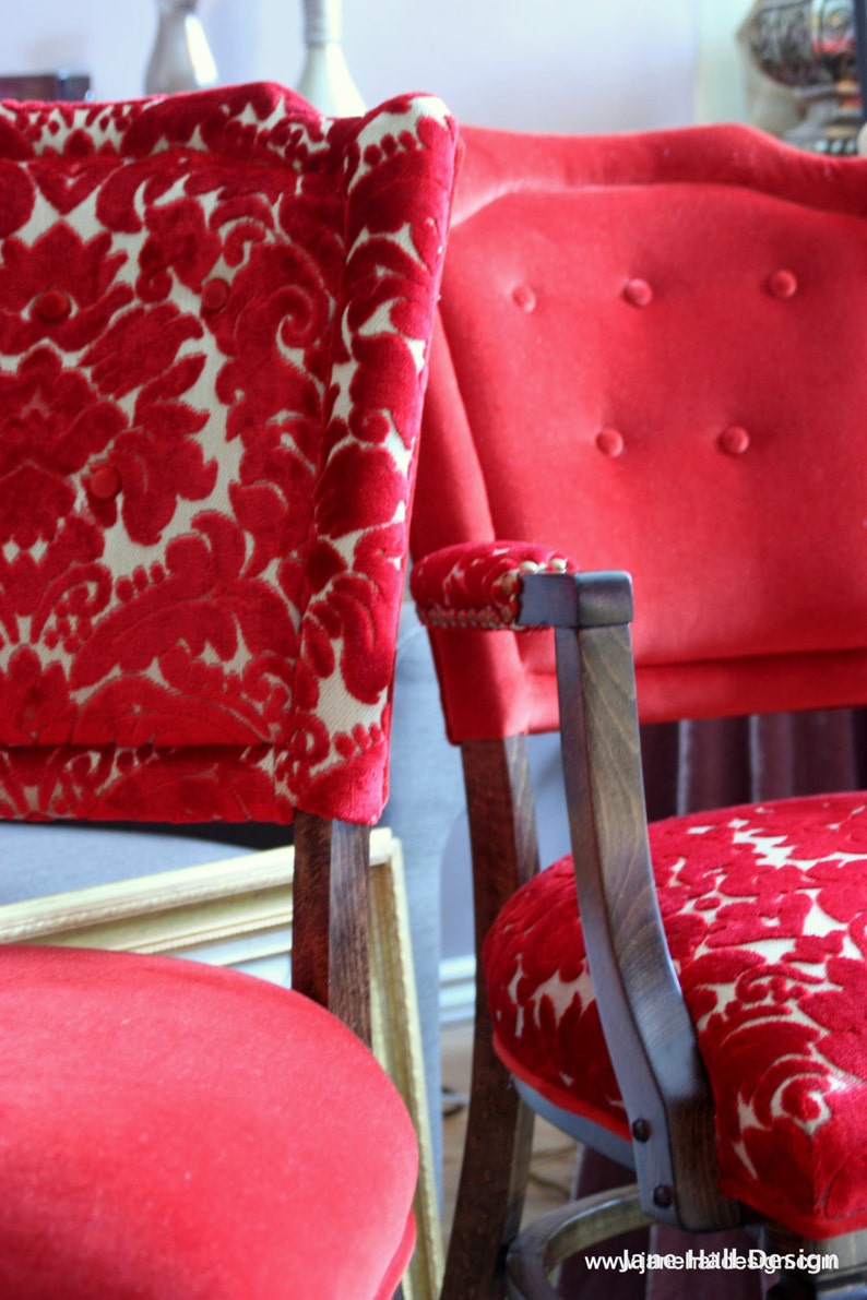 May include: Two red velvet chairs with ornate floral patterns. The chairs have wooden frames and are upholstered in a rich red fabric. The chairs are in a room with a light gray wall and a wooden floor.