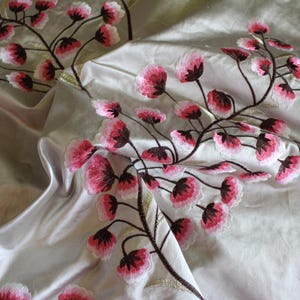 May include: Close-up of a fabric with embroidered floral branches. The flowers are pink and white with dark centers, set against a pale, satiny background. The branches are brown with gold accents, creating a delicate, nature-inspired design.