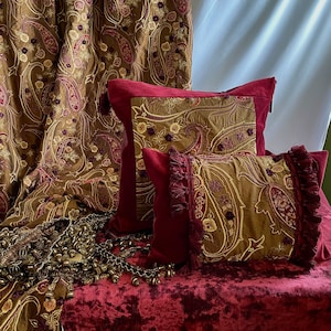 May include: A collection of home textiles with a paisley design. The image shows a curtain, two cushions, and decorative trim. The colour scheme is burgundy, gold, and brown, creating a luxurious aesthetic for interior design.