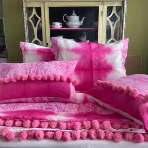 May include: A bed set with a striking pink and white tie-dye design. The set includes pillows with pom-pom edging and a matching duvet cover. The bedding is arranged on a white surface, creating a bold bedroom aesthetic. The pillows and duvet are in cm.