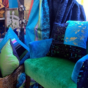 Op de afbeelding: Een tealblauwe fluwelen fauteuil met een groene fluwelen zitting. De stoel is versierd met een patchwork kussen met zwarte en gouden geometrische patronen en een tealblauw en groen bloemenpatroon. De stoel staat voor een blauw en teal gepatroneerd stof.