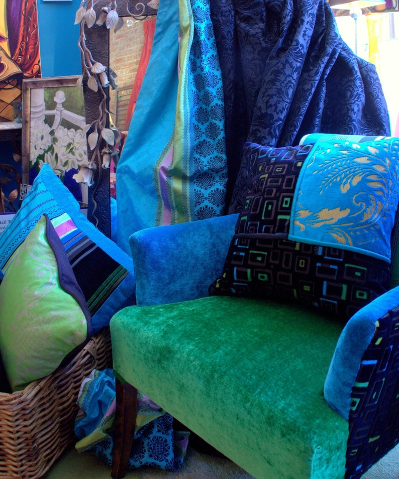 Op de afbeelding: Een tealblauwe fluwelen fauteuil met een groene fluwelen zitting. De stoel is versierd met een patchwork kussen met zwarte en gouden geometrische patronen en een tealblauw en groen bloemenpatroon. De stoel staat voor een blauw en teal gepatroneerd stof.