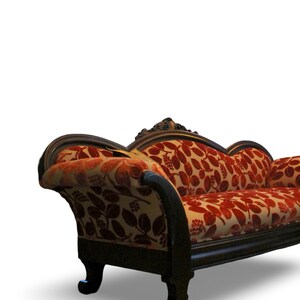 May include: An antique chaise lounge with a dark wooden frame and legs. The seat and back are upholstered in a cream-colored fabric with a red floral pattern. The chaise lounge has a curved armrest and back.