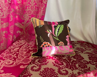 May include: A decorative pillow with a patchwork design featuring floral and striped patterns in shades of brown, pink, and green. The pillow has a yellow-green trim and is set against a backdrop of pink floral fabric.