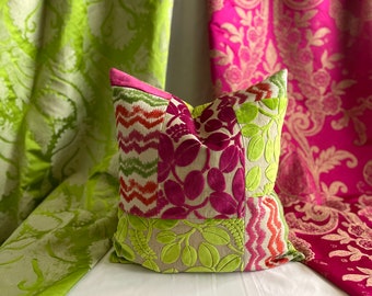 May include: A decorative pillow with a patchwork design featuring various patterns and colors. The pillow has sections of green, pink, and beige fabrics with leaf and zigzag patterns. The pillow is set against a backdrop of green and pink patterned curtains.