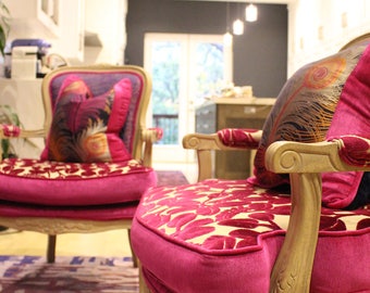 May include: Two ornate armchairs with a vibrant fuchsia hue. The chairs feature carved wooden frames and plush cushions with a floral pattern in red and gold. Pillows with feather designs add a touch of elegance. The chairs sit on a patterned rug.