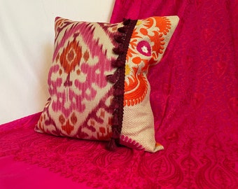 May include: A decorative pillow with a pink, orange, and white ikat pattern on one side and a floral embroidery design on the other side. The pillow has a maroon tassel trim.
