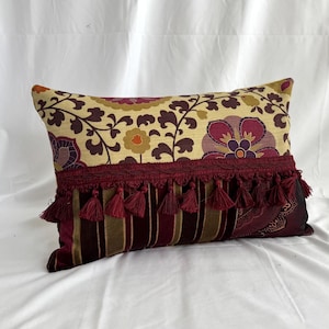 May include: Rectangular cushion with a floral pattern in cream, burgundy, and purple. The cushion features a burgundy tassel trim and a striped and patterned bottom section. Ideal for home decor.