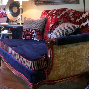 May include: A blue velvet sofa with gold and red trim. The sofa has two pillows, one with a red and gold floral pattern and the other with a striped pattern. The sofa is sitting in a room with a wooden floor.