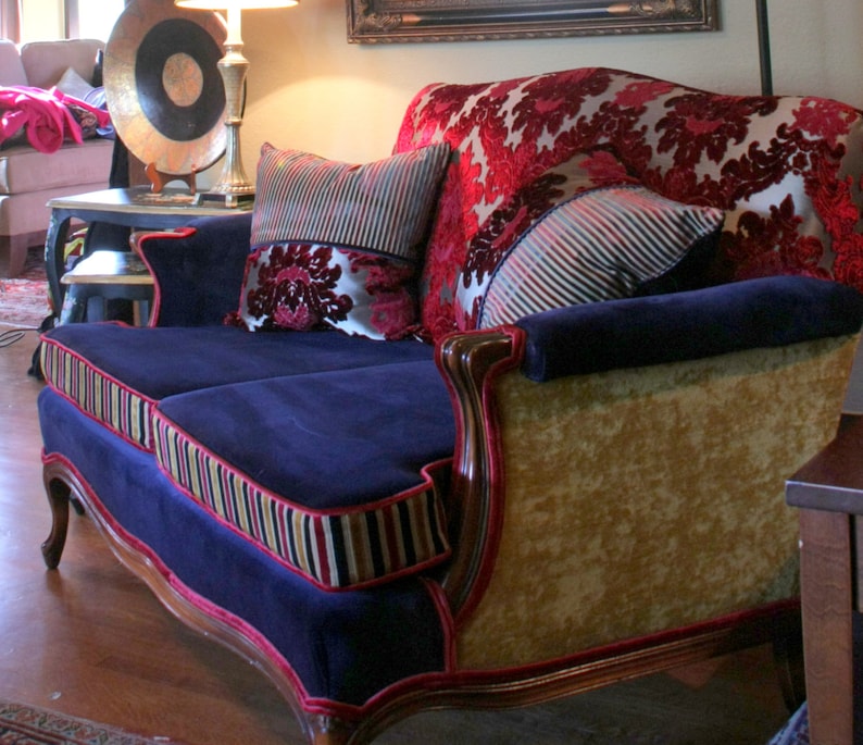 May include: A blue velvet sofa with gold and red trim. The sofa has two pillows, one with a red and gold floral pattern and the other with a striped pattern. The sofa is sitting in a room with a wooden floor.