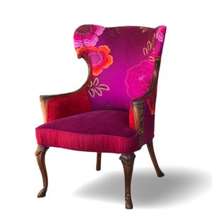 May include: A decorative wingback chair with a high back and curved arms. The chair features a vibrant floral pattern in shades of pink, orange, and purple on the back and arms. The seat is a solid deep pink, and the wooden legs are dark brown.