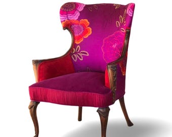 May include: A decorative wingback chair with a high back and curved arms. The chair features a vibrant floral pattern in shades of pink, orange, and purple on the back and arms. The seat is a solid deep pink, and the wooden legs are dark brown.
