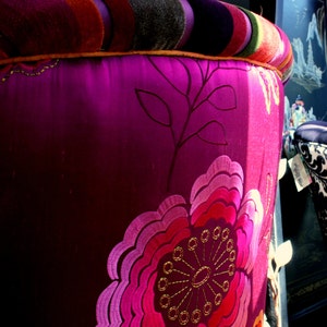 May include: A close-up of a decorative stool with a vibrant, floral design. The stool features a deep magenta fabric with an embroidered flower in shades of pink and red. The top is striped with orange, purple, and green.