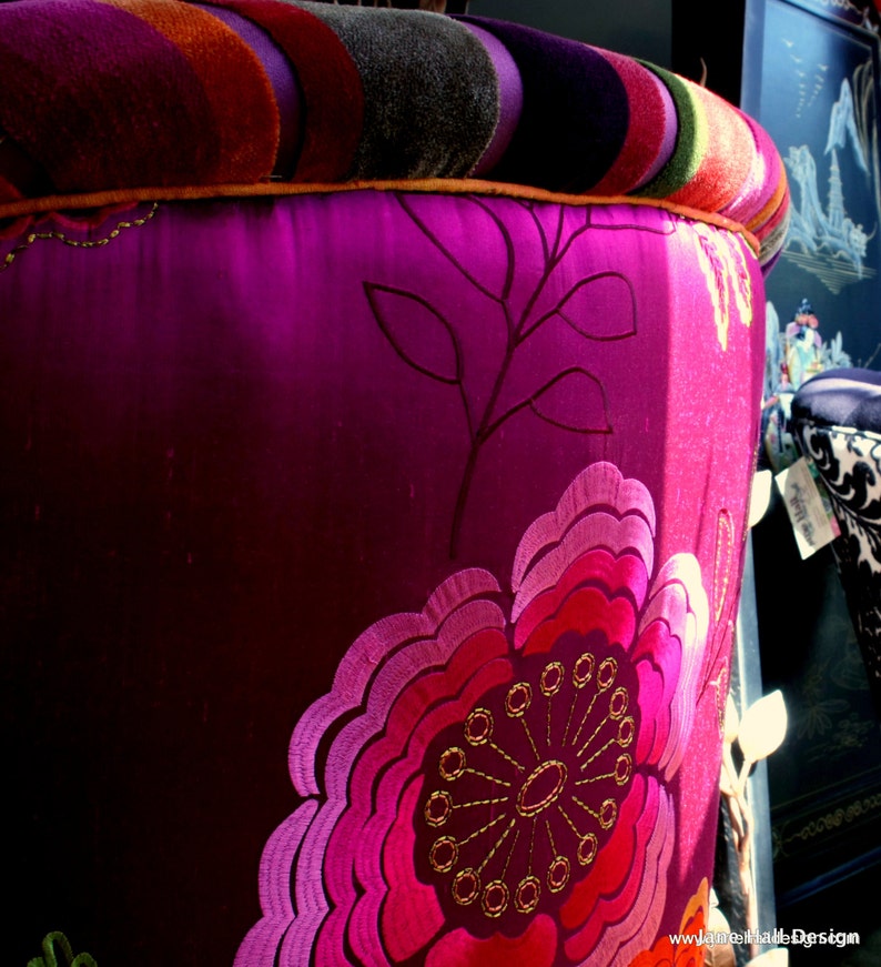 May include: A close-up of a decorative stool with a vibrant, floral design. The stool features a deep magenta fabric with an embroidered flower in shades of pink and red. The top is striped with orange, purple, and green.