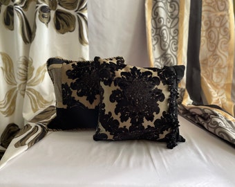 May include: Two decorative throw pillows with black velvet fabric and gold embroidery. The pillows have a floral pattern and are accented with black tassels.