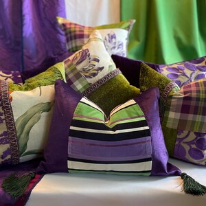 May include: A collection of decorative throw pillows in various patterns and colors. Pillows feature floral, plaid, and striped designs in shades of purple, green, and white. Some pillows have decorative trim and tassels.