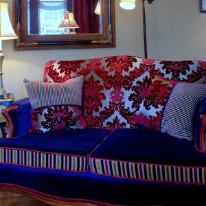 May include: A blue velvet sofa with a red and gold patterned upholstery. The sofa has two pillows, one with a striped pattern and the other with a floral pattern. The sofa is in a living room setting.